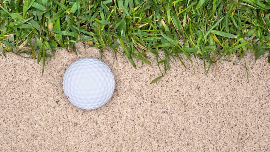A golf ball in a sand trap next to the rough highlights the mistakes one can make playing the game