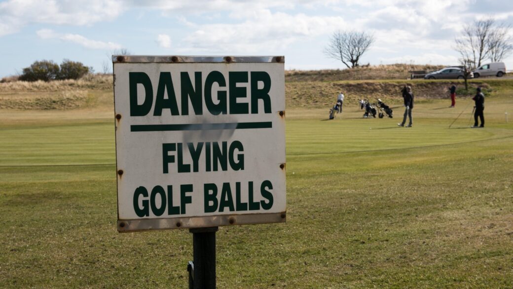 Warning sign reading "Danger Flying Golf Balls" on a golf course, with golfers and caddies in the background under a partly cloudy sky